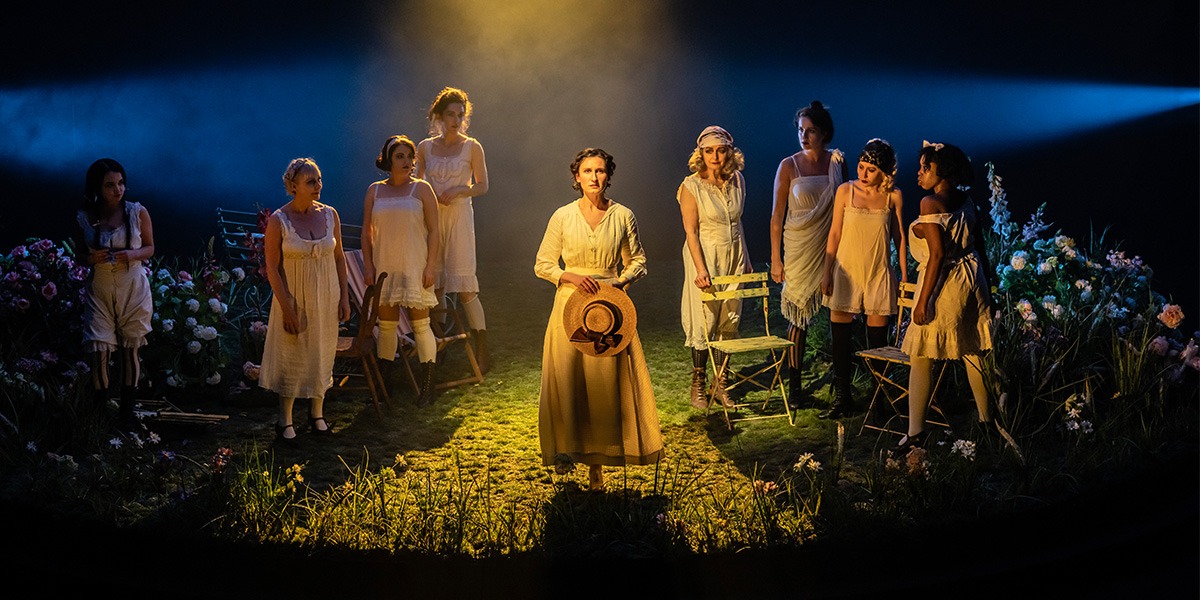 The company of Mrs Warrens Profession stand on stage upon green grass whilst surrounded by wild flowers.