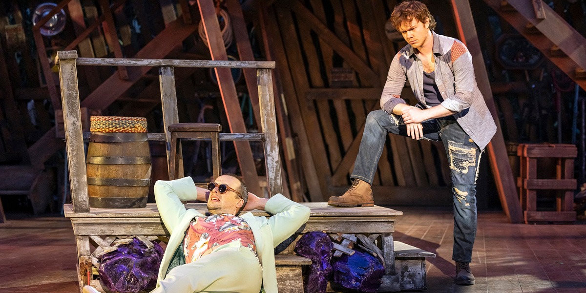 Two actors are on stage, with one seated on a lavish chair and the other standing beside a wooden crate, set against a rustic backdrop.