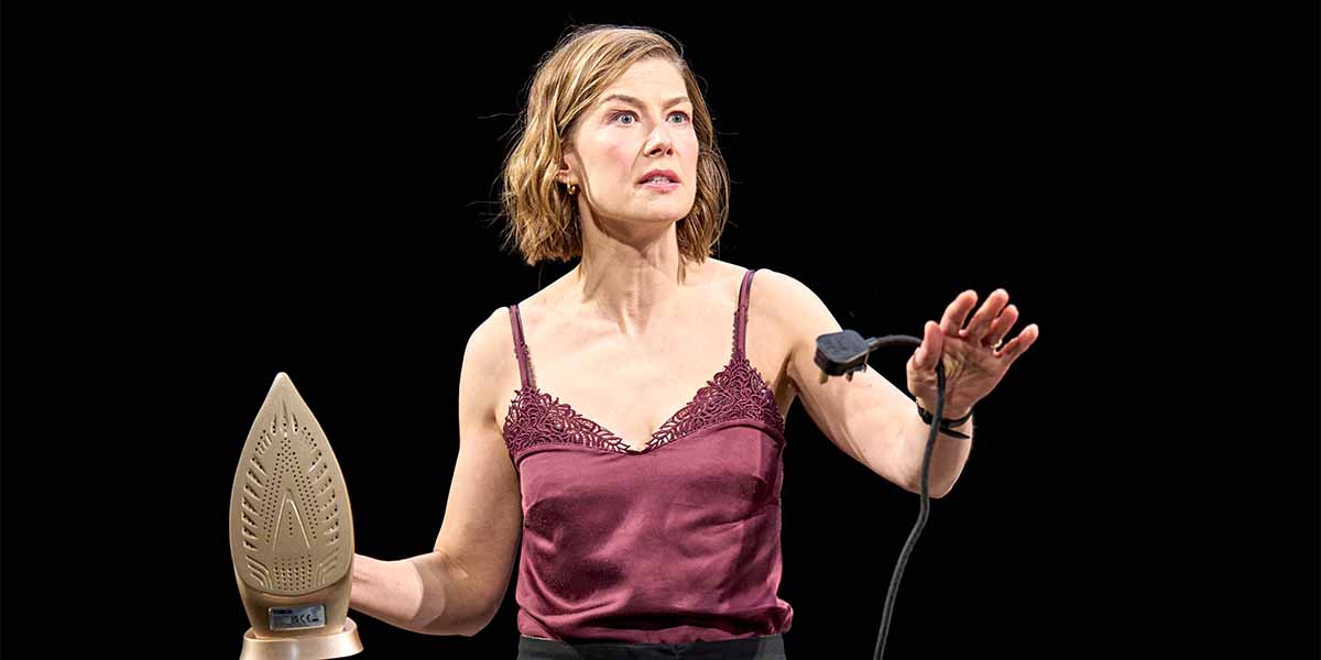 Rosamund Pike, in a burgundy top, gestures with an iron in hand against a black background.