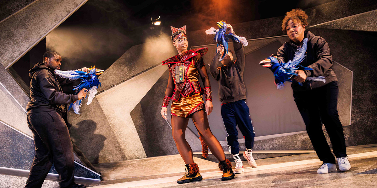 Four performers in a theatrical setting, one wearing a vibrant traditional outfit and the others in casual wear, engaging dynamically with colorful fabrics on a dramatically lit stage with geometric designs.