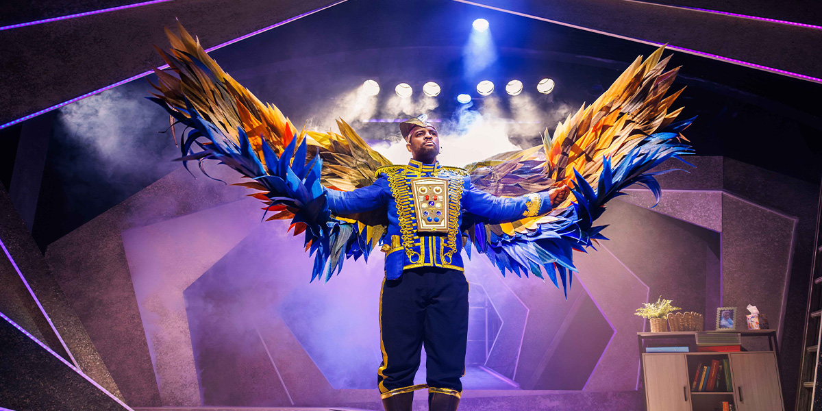 Performer on stage wearing an elaborate costume with large, colorful feathered wings, illuminated by stage lights.