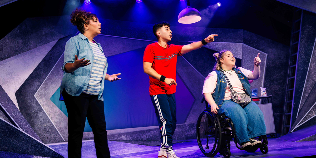 Three actors on stage in a theatrical production, expressing excitement and engagement in their scene. The setting includes vibrant lighting and a modern, geometric backdrop.