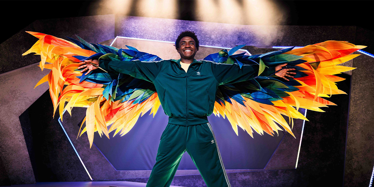Person in a green tracksuit with expansive, colorful feathered wings, standing on a stage with a purple backdrop.