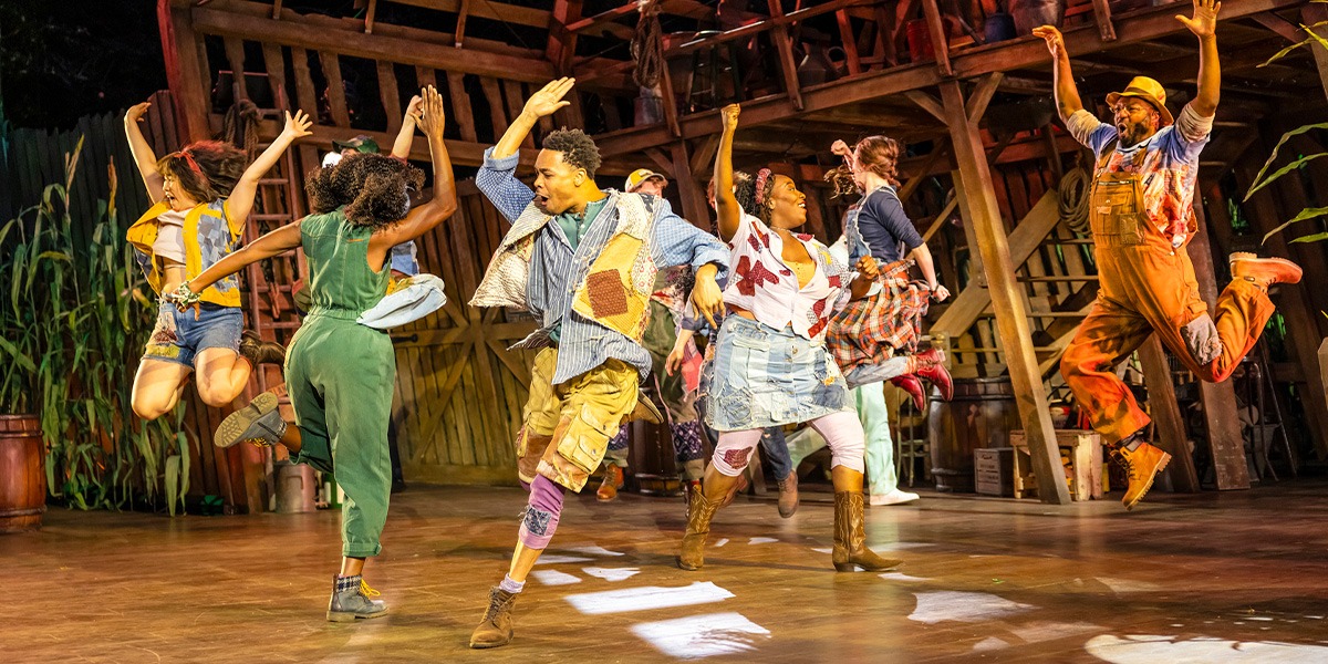 The company of Shucked are wearing colourful attire with patches sewn on. They are mid stage, leaping into the air.