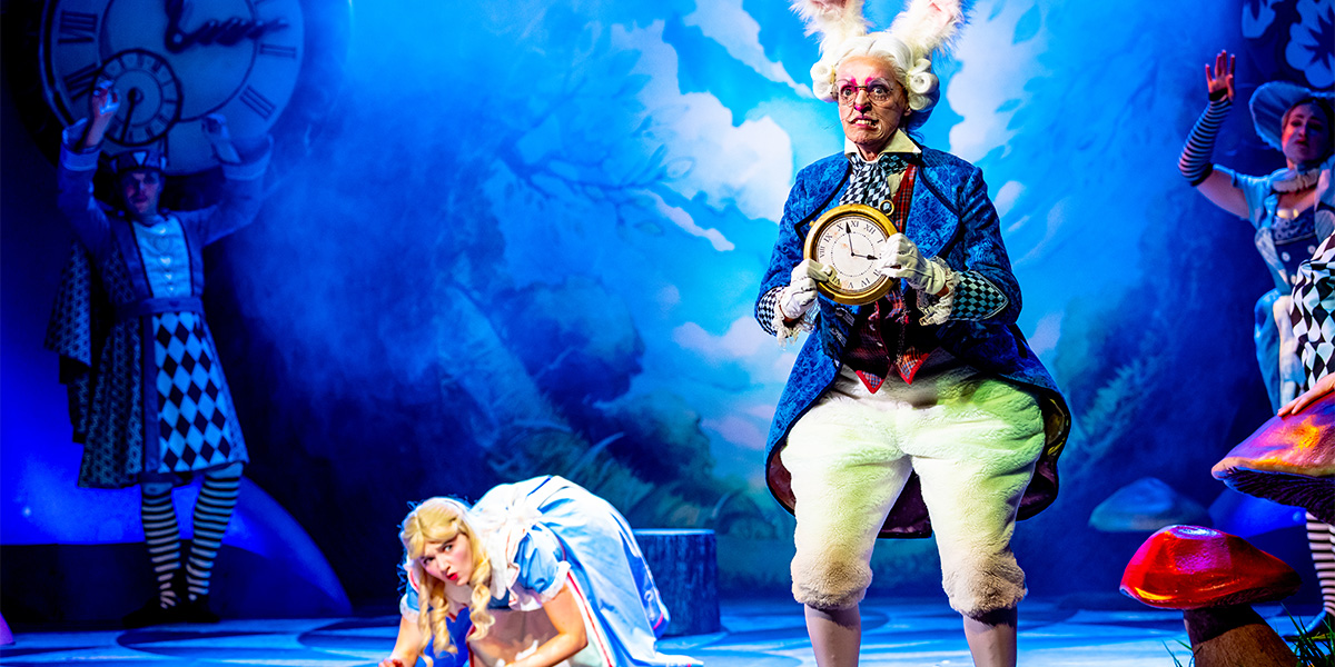 A scene from "Alice in Wonderland" showing a person dressed as the White Rabbit holding a large clock, with Alice in the background. The setting has whimsical decorations and a vibrant, colourful backdrop resembling a forest.