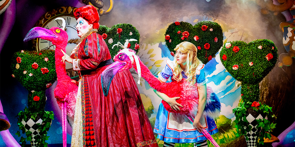 Two characters from "Alice in Wonderland," the Queen of Hearts and Alice, engage in a scene on stage with vividly decorative props including flamingo statues and roses.