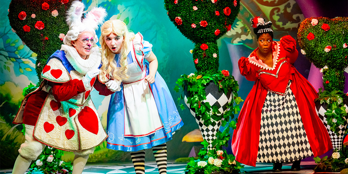 Scene from 'Alice in Wonderland' showing three characters on stage: the White Rabbit and Alice in the left half, and the Queen of Hearts on the right, all in colorful costumes amid a vibrant set design with oversized flowers and card motifs.