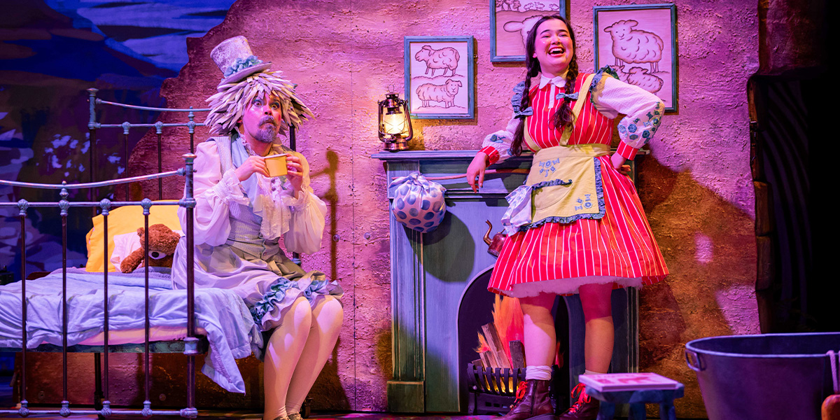 Two performers in colorful costumes on stage, one seated with a cup and another standing beside a fireplace, in a whimsically decorated room resembling a children's story setting.