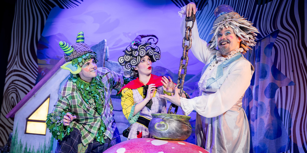 Three actors on stage in a theatrical production interact playfully around a large, colourful prop.