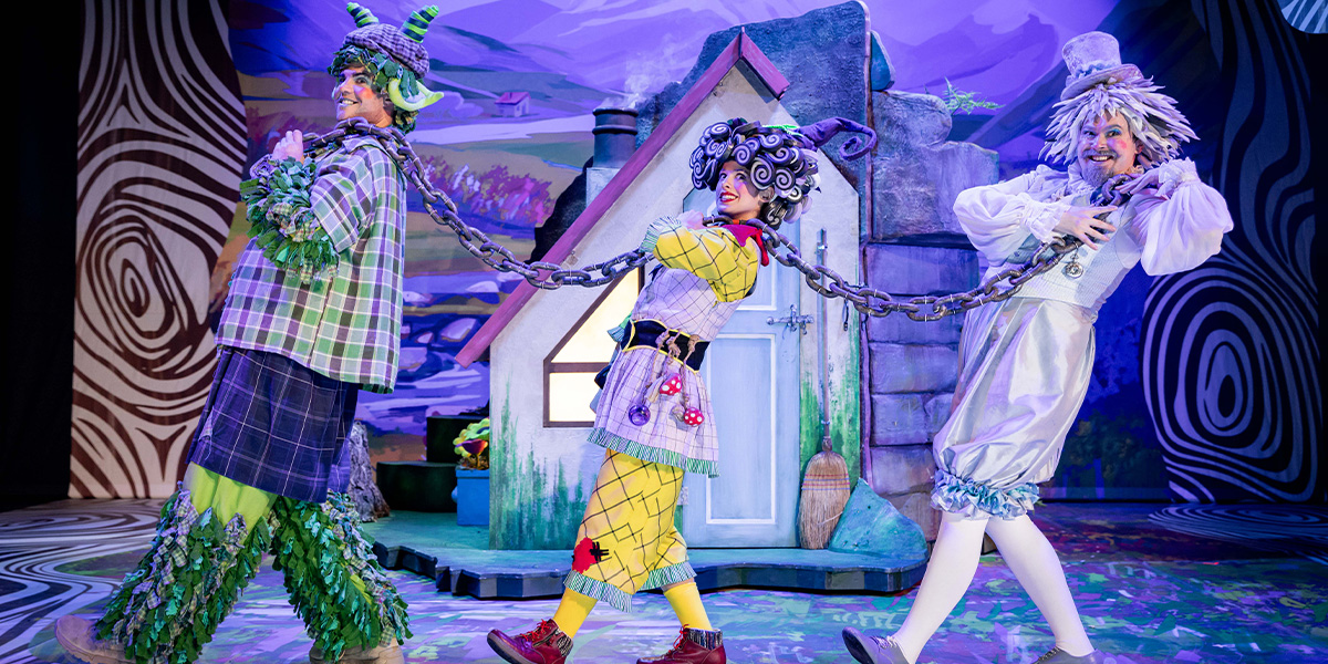 Three actors in colourful costumes perform on stage in a whimsical setting, depicting a scene with playful and exaggerated expressions. One is dressed in green with foliage-like elements, another in a patchwork outfit, and the third in white with fluffy details, all linked by a chain near a stylized cottage backdrop.