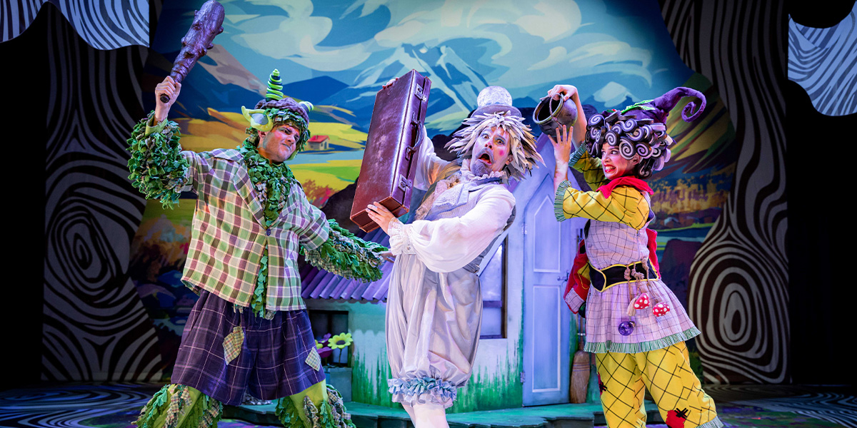 Three actors in colourful, whimsical costumes perform on stage in a theatrical production, each holding props like a branch and a suitcase, set against a stylized backdrop.