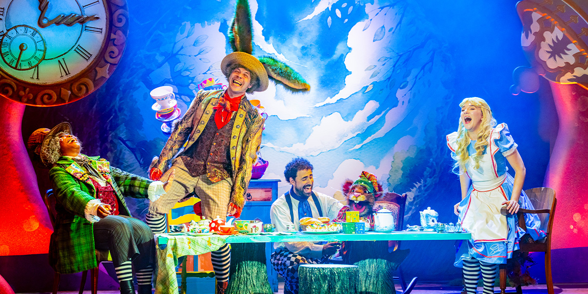 Scene from 'Alice in Wonderland' showing characters at the Mad Hatter's tea party, seated around a colorful table with various teacups and pots under a whimsical, large clock and sky-themed backdrop.