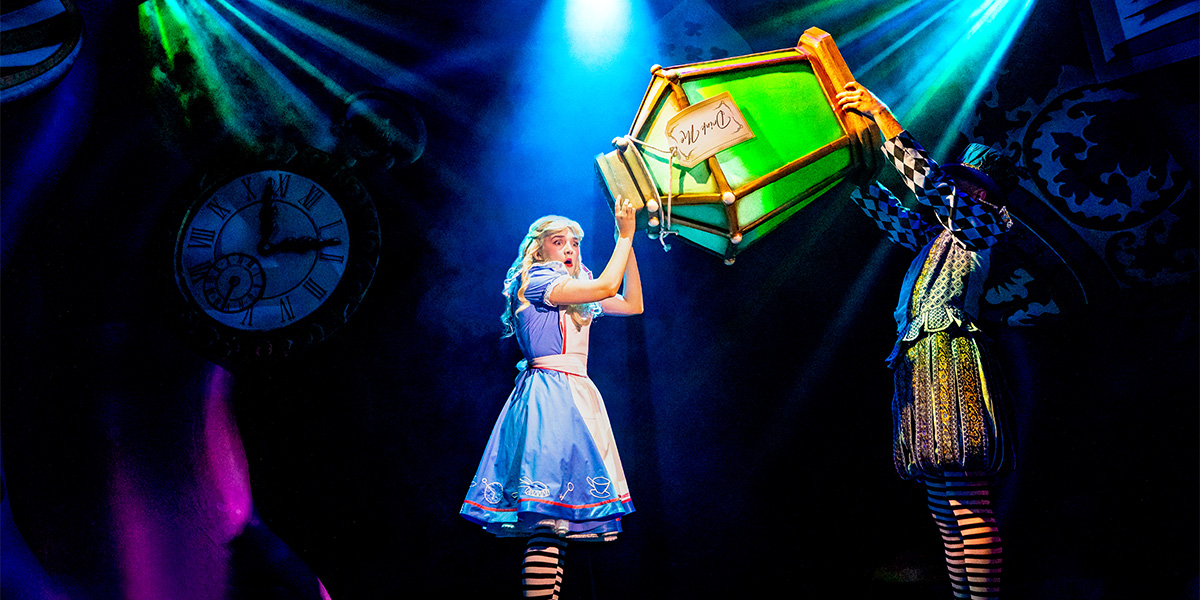 A performer on stage interacting with a large, illuminated book prop in a theatrical setting with colorful lighting and whimsical backdrop featuring a clock and tree silhouettes, in a scene from the musical "Alice in Wonderland."