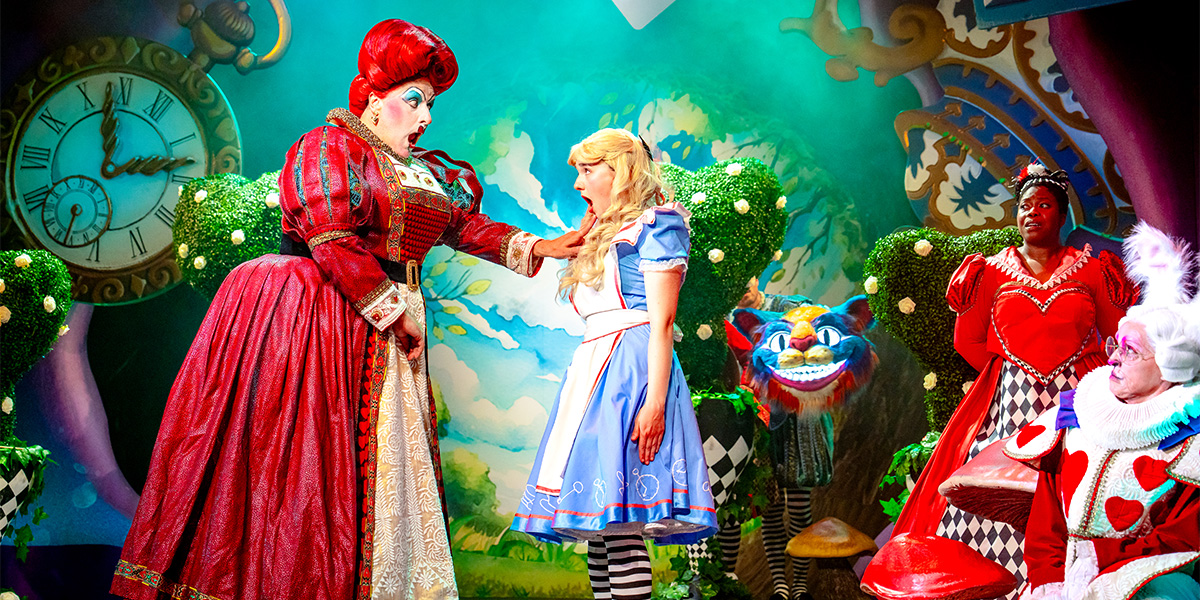 A vibrant theatrical scene from "Alice in Wonderland" featuring characters Alice, the Red Queen, and another character in a colorful, whimsical stage setting with oversized props like a clock and flora.