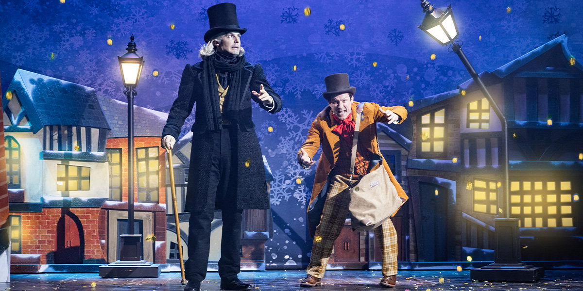Daniel Fraser and Matt Cavendish pose together, smiling, in festive costumes against a Christmas-themed backdrop.