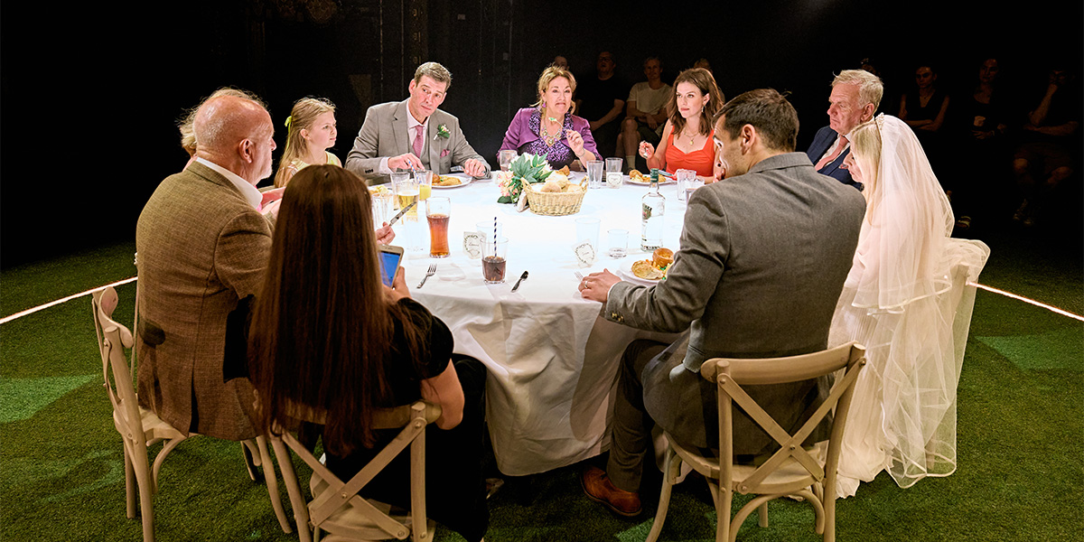 A group of people seated around a rectangular dinner table in a theatrical setting, featuring a person in a wedding dress and another in a suit at the head of the table.
