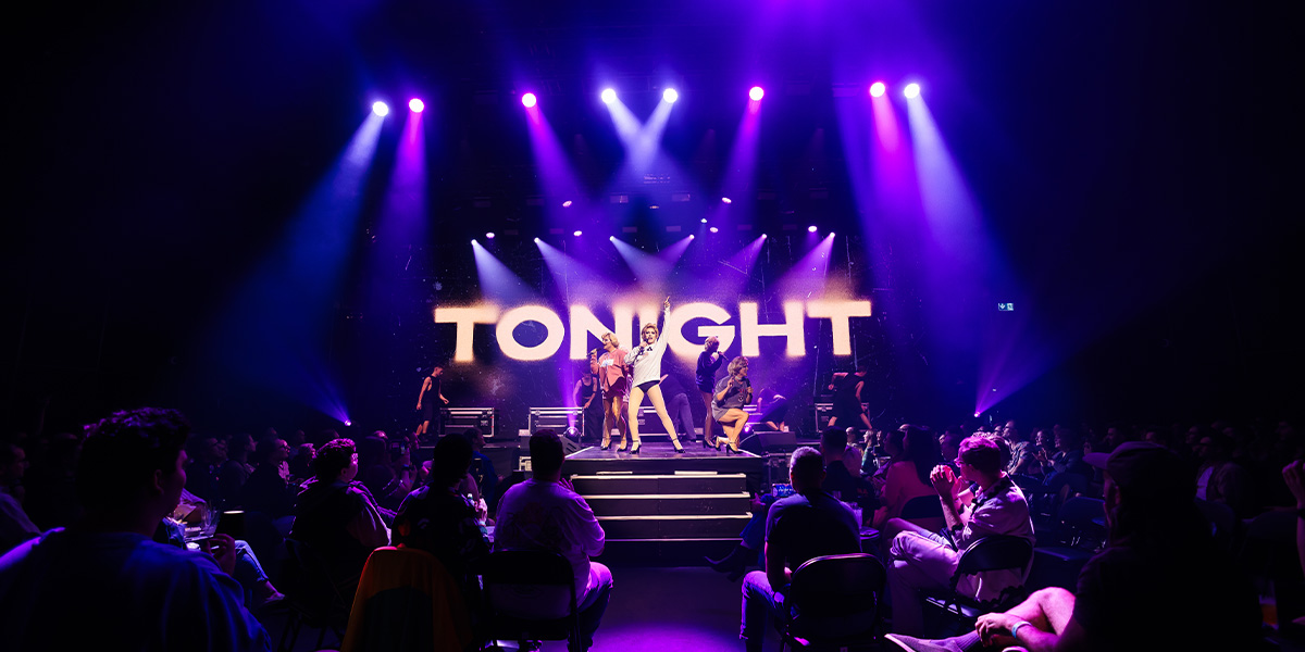 Performers on stage at a live concert with bright lights and 'TONIGHT' displayed in the background, with an audience in the foreground.