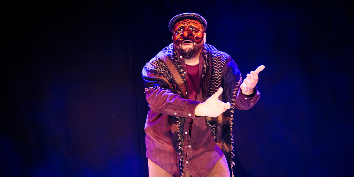 A performer wearing a traditional mask and shawl gestures expressively against a dark background.