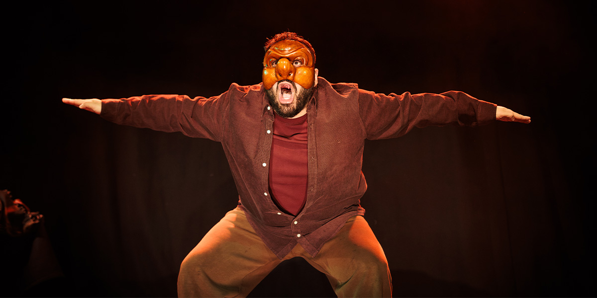 A performer in a brown shirt and mask poses dramatically with outstretched arms against a dark background.