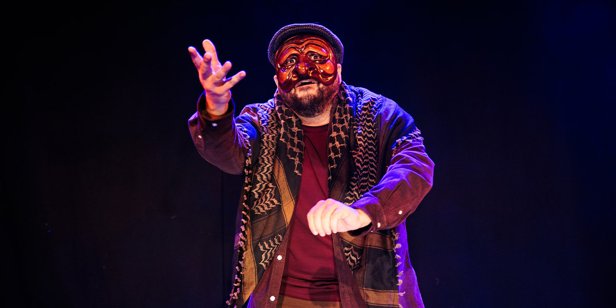 A performer in a red mask and patterned shawl gestures expressively against a dark background.