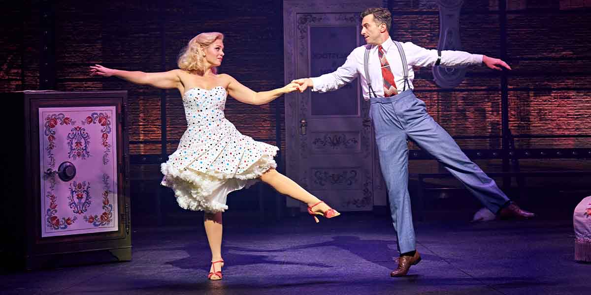 Joanna Woodward and Marc Antolin perform a dance, holding hands, in vibrant costumes on stage.