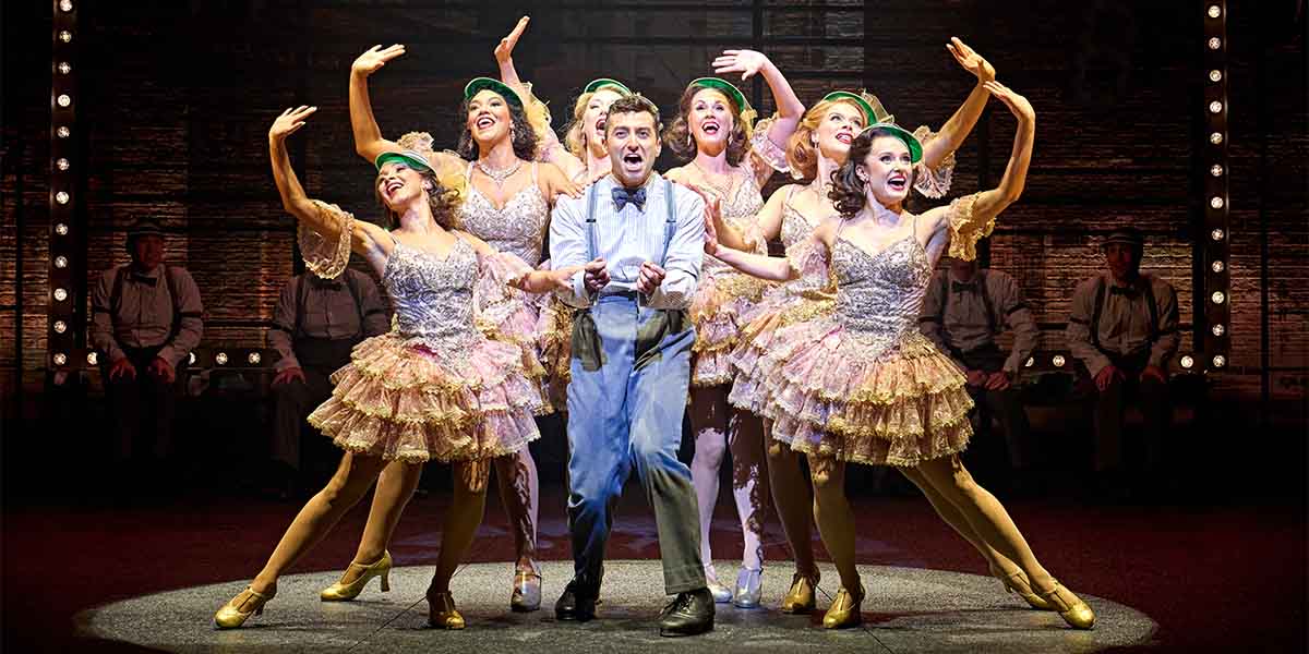 Marc Antolin as Leo Bloom performs centre stage with six dancers in glittery costumes, all smiling.