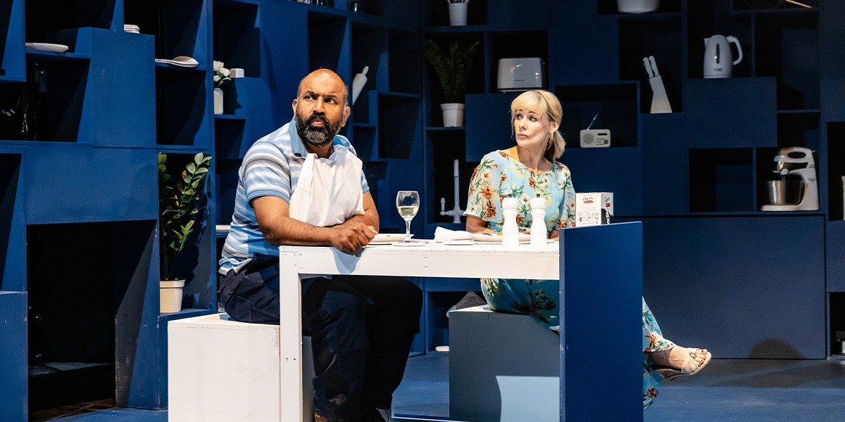 Two actors are on stage seated at a table, with a backdrop of blue shelves filled with various household items.