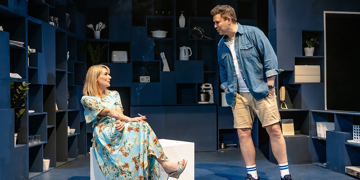 Two actors are on stage, one sitting and the other standing, with a modern set design featuring multiple shelves and household items.