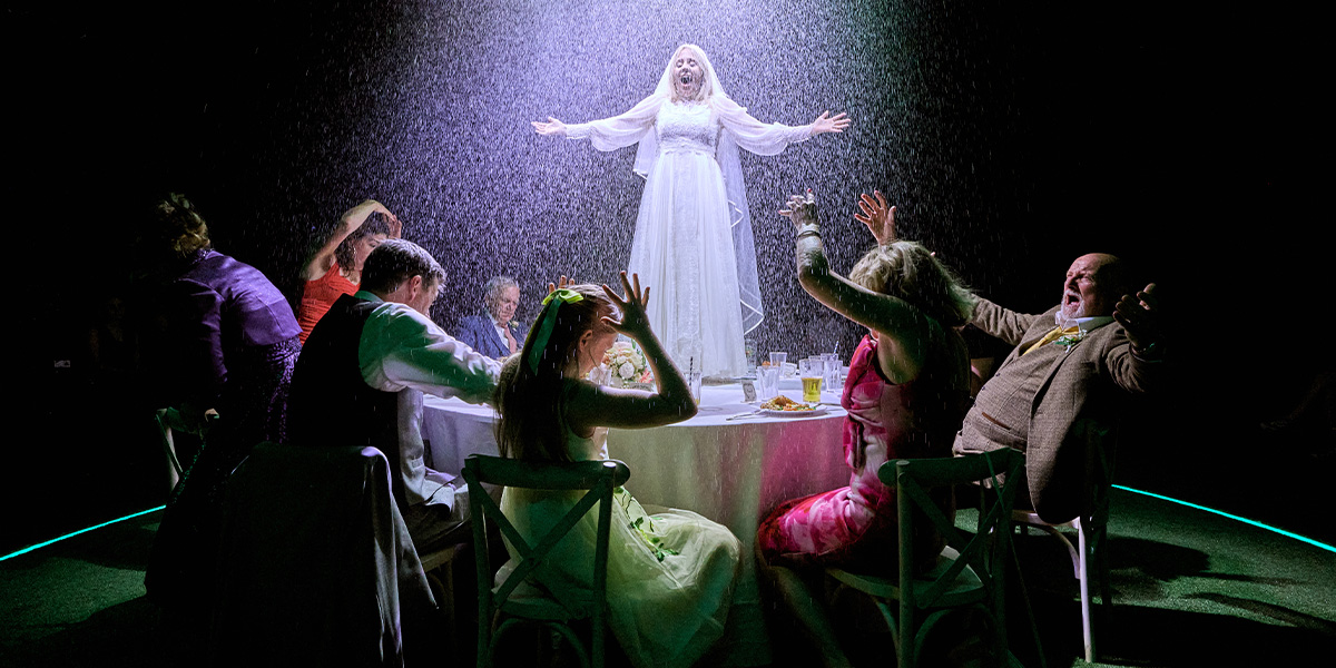 A theatrical scene from a performance, featuring a person in a white costume standing with outstretched arms on a table surrounded by other individuals in festive attire, reacting dramatically under artificial rain and dramatic lighting.