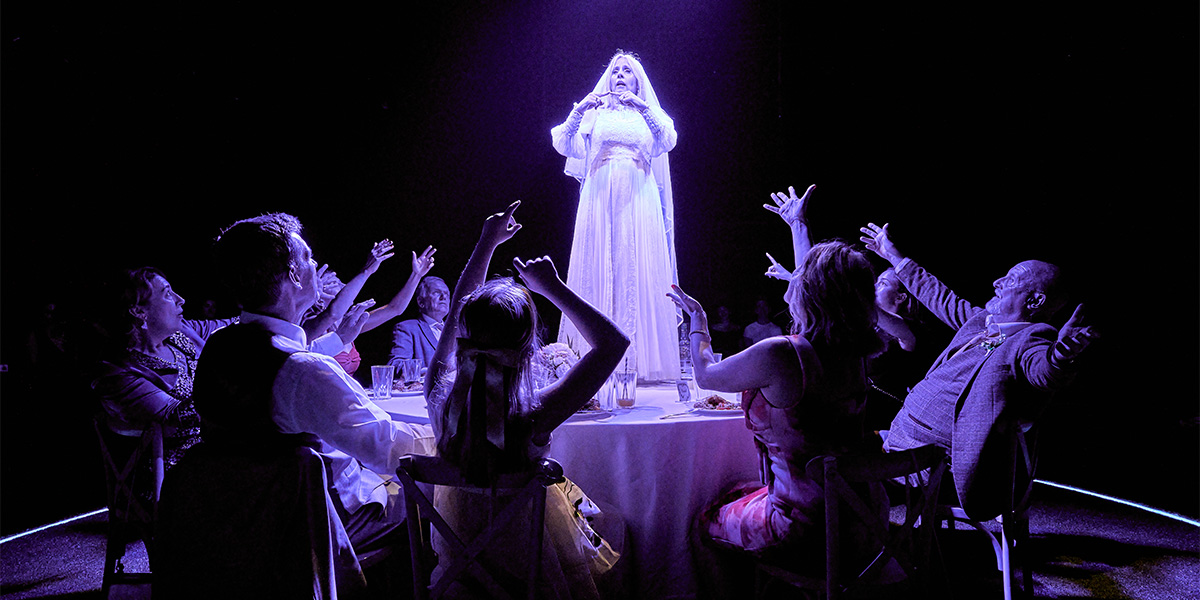 A performer in a white dress stands under a spotlight on a stage, singing to an audience seated around the stage, who are clapping and reaching towards the performer.