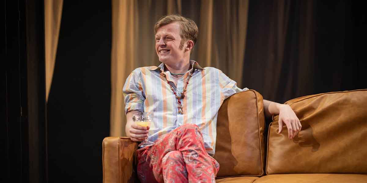 William Robinson sits on a brown sofa, smiling, wearing a striped shirt and patterned trousers, holding a drink.