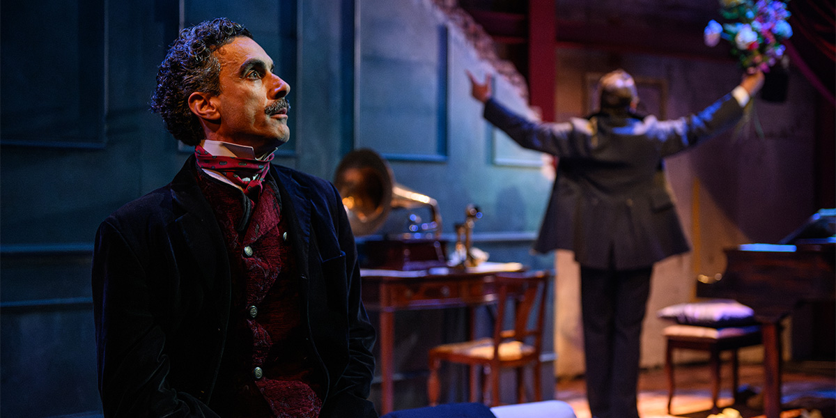 A person in a historical costume, featuring a dark suit and red cravat, looks thoughtfully to the side on a stage set resembling a 19th-century room, with another individual in the background holding up a bunch of flowers.