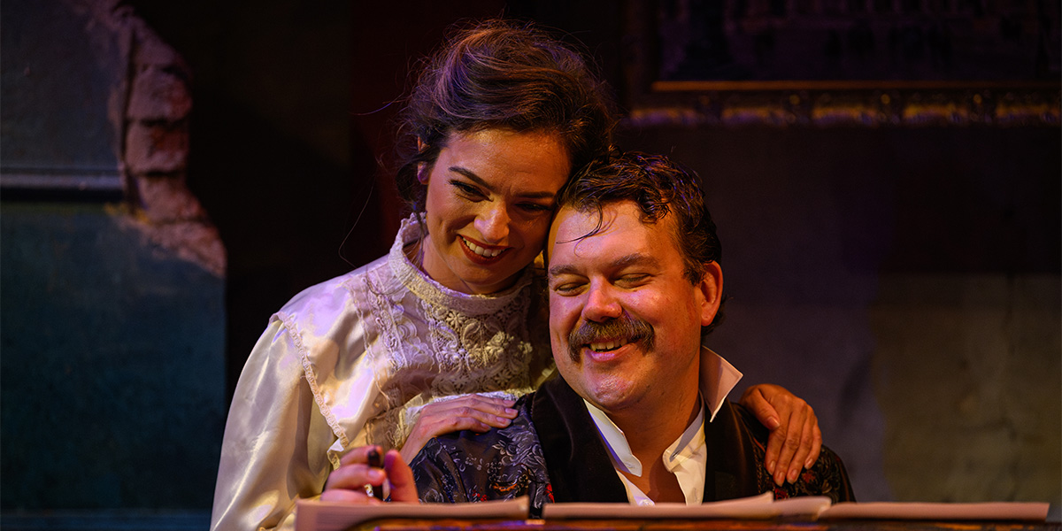 Two actors on stage during a theatrical performance, one standing and smiling while the other sits and laughs, both dressed in period costumes.