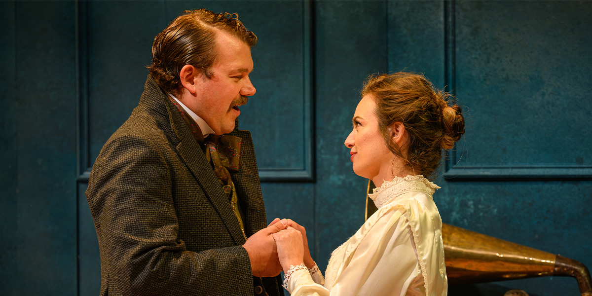 Two actors performing a scene on stage, one in a brown suit and the other in a white period dress, engaging warmly with each other in a set designed to look like a vintage room with blue walls.