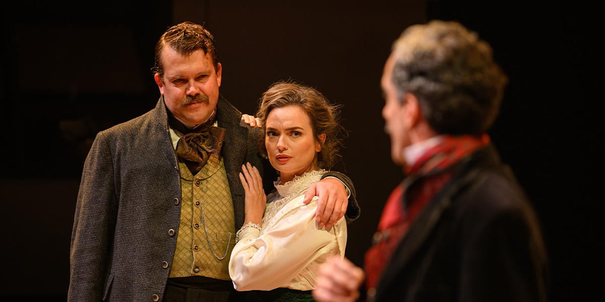 Three actors in period costume on stage in a dramatic scene, featuring a central figure looking concerned, flanked by another who is comforting them, and a third character observing intently.