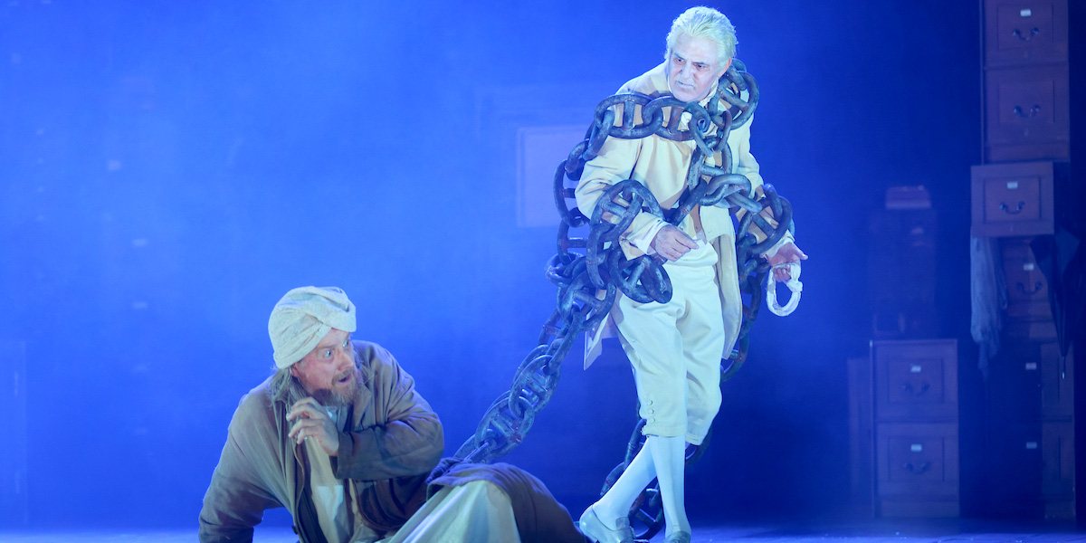 A ghostly figure in chains confronts a terrified man in a dimly lit, atmospheric stage setting.