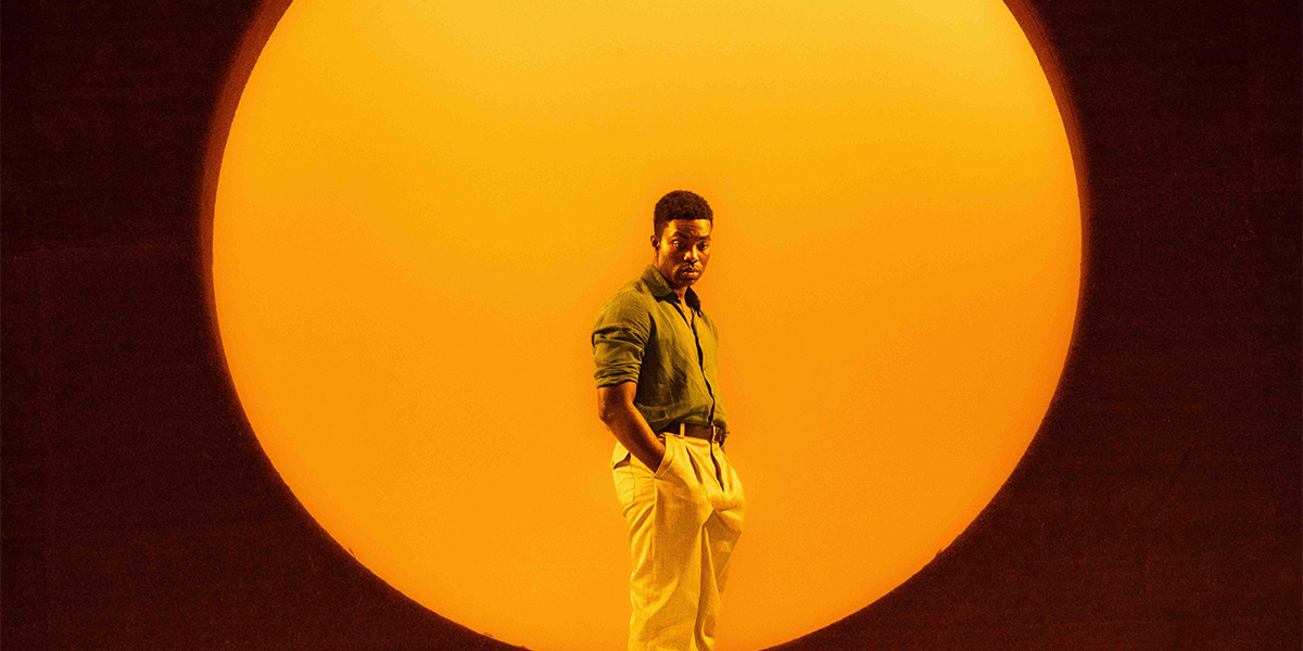 Paapa Essiedu as Chris Keller stands confidently against a large orange backdrop, wearing a green shirt and beige trousers.