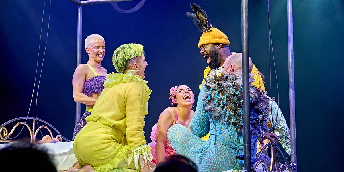 Four actors in vibrant costumes enjoying a lively scene on a theatrical stage, featuring imaginative outfits in yellow, green, and blue.