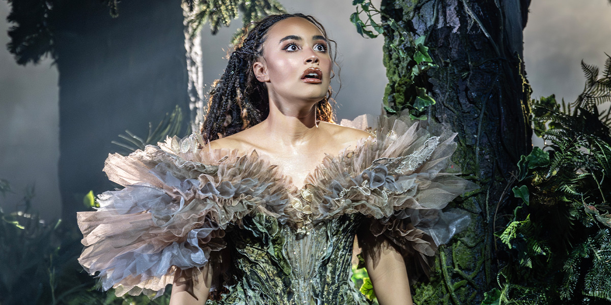 A person wearing an elaborate green dress stands surrounded by trees.