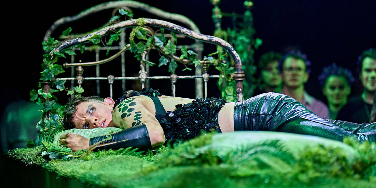 Performer in a black and green costume lies dramatically on a grass covered bed with other performers watching in the background.