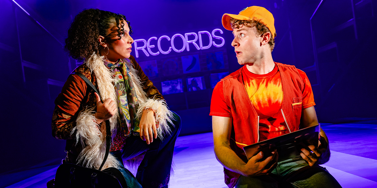 Three dancers in vibrant costumes perform energetically on stage, with colourful lights illuminating the background.