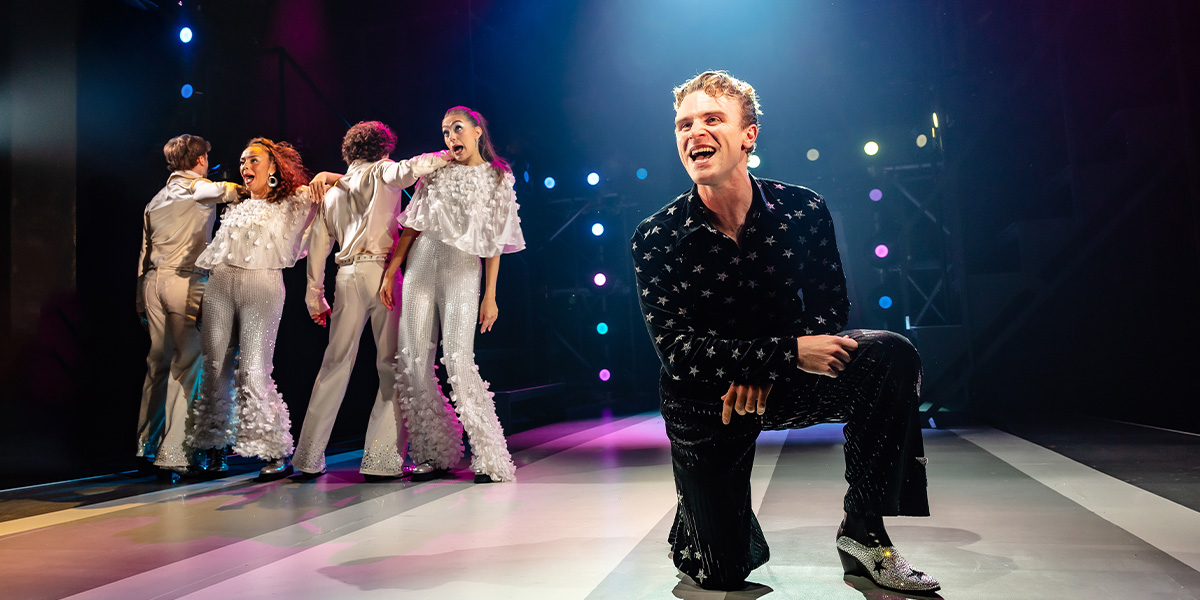 A diverse group of dancers in vibrant costumes performs energetically on stage, with colourful lighting in the background.