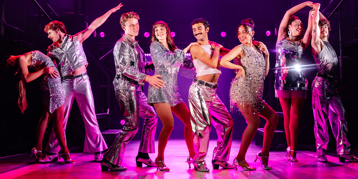 A diverse group of dancers in vibrant costumes performs energetically on stage, illuminated by colourful lights.