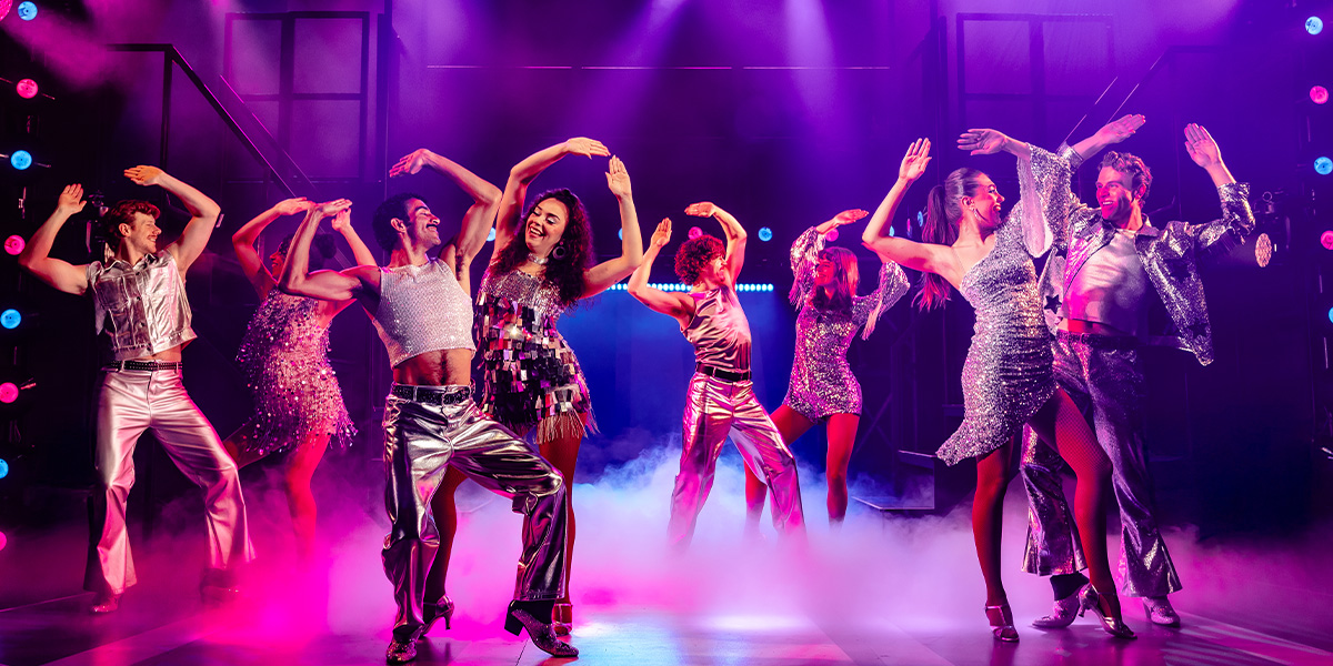 A diverse group of performers in vibrant costumes, energetically dancing on stage with colourful lighting in the background.