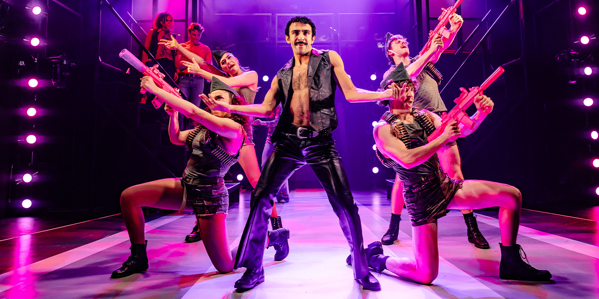 A diverse group of performers in vibrant costumes dance energetically on stage, with colourful lighting in the background.