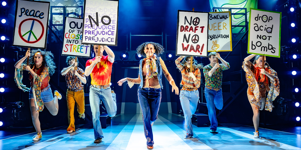 A diverse group of performers in vibrant costumes poses energetically on stage, with colourful lights in the background.