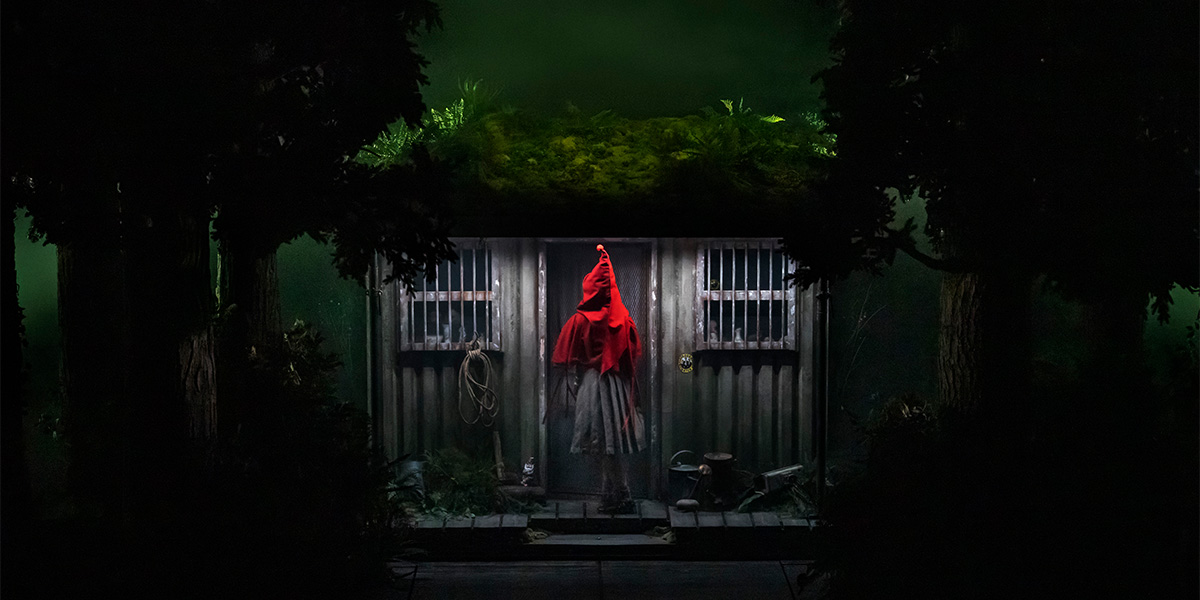 A person wearing a red cape stands outside a cottage in the woods, about to enter.
