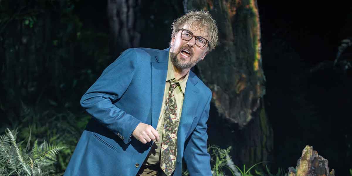 John Owen-Jones performs in a blue suit with a patterned tie, surrounded by a forest backdrop.