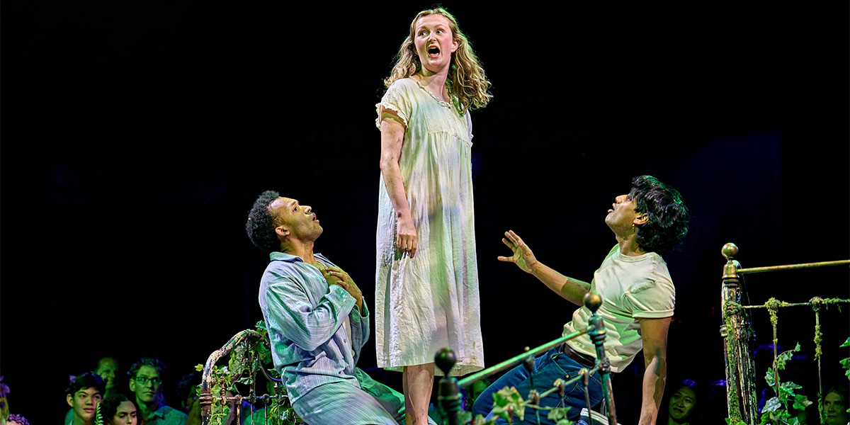 Three actors perform on stage in a theatrical production, one standing and expressing dramatically while the other two sit and look up, engaged in the scene.