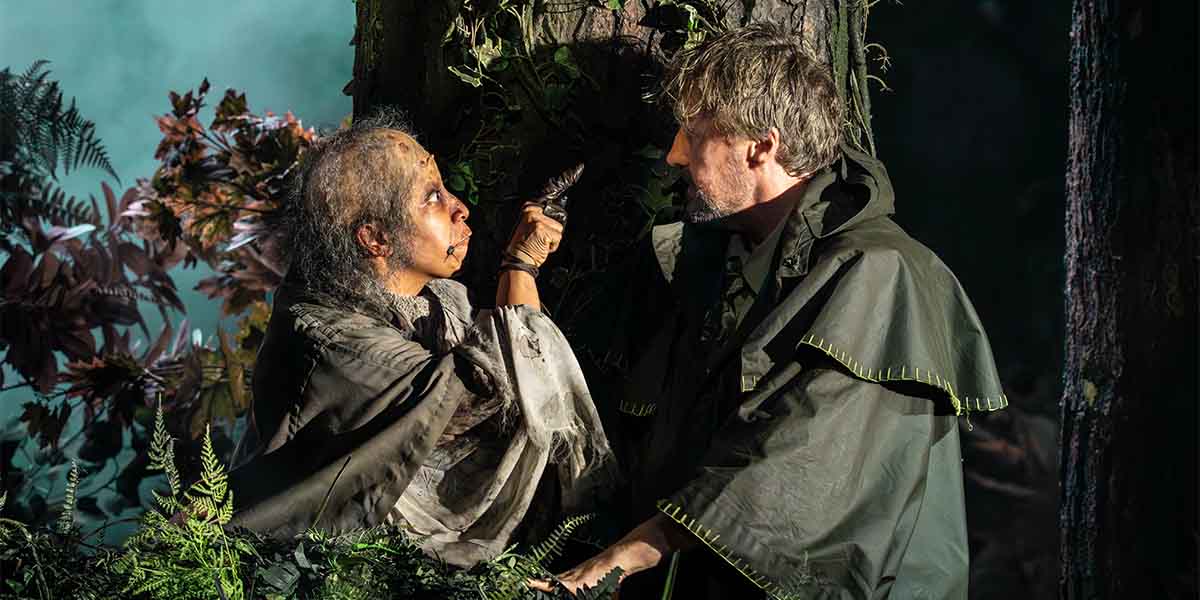 Melanie La Barrie and John Owen-Jones interact dramatically in a forest setting, surrounded by foliage.